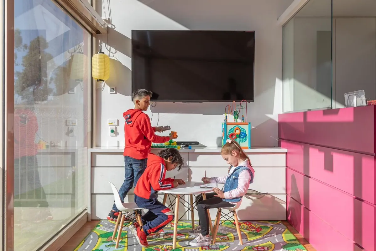 Three young children drawing at a small table in a bright children's area, with toys and an activity cube nearby.