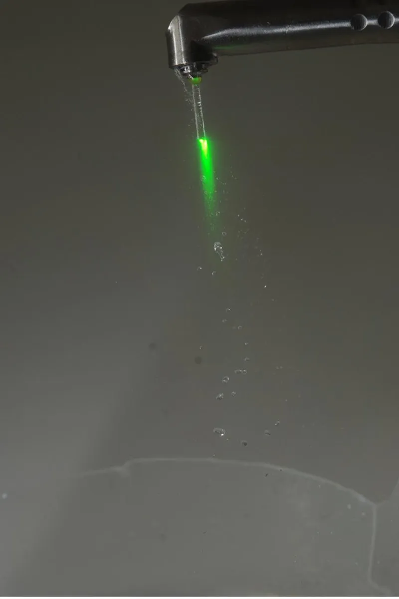 Photograph of a dental laser handpiece with a green guide light shining through falling water droplets